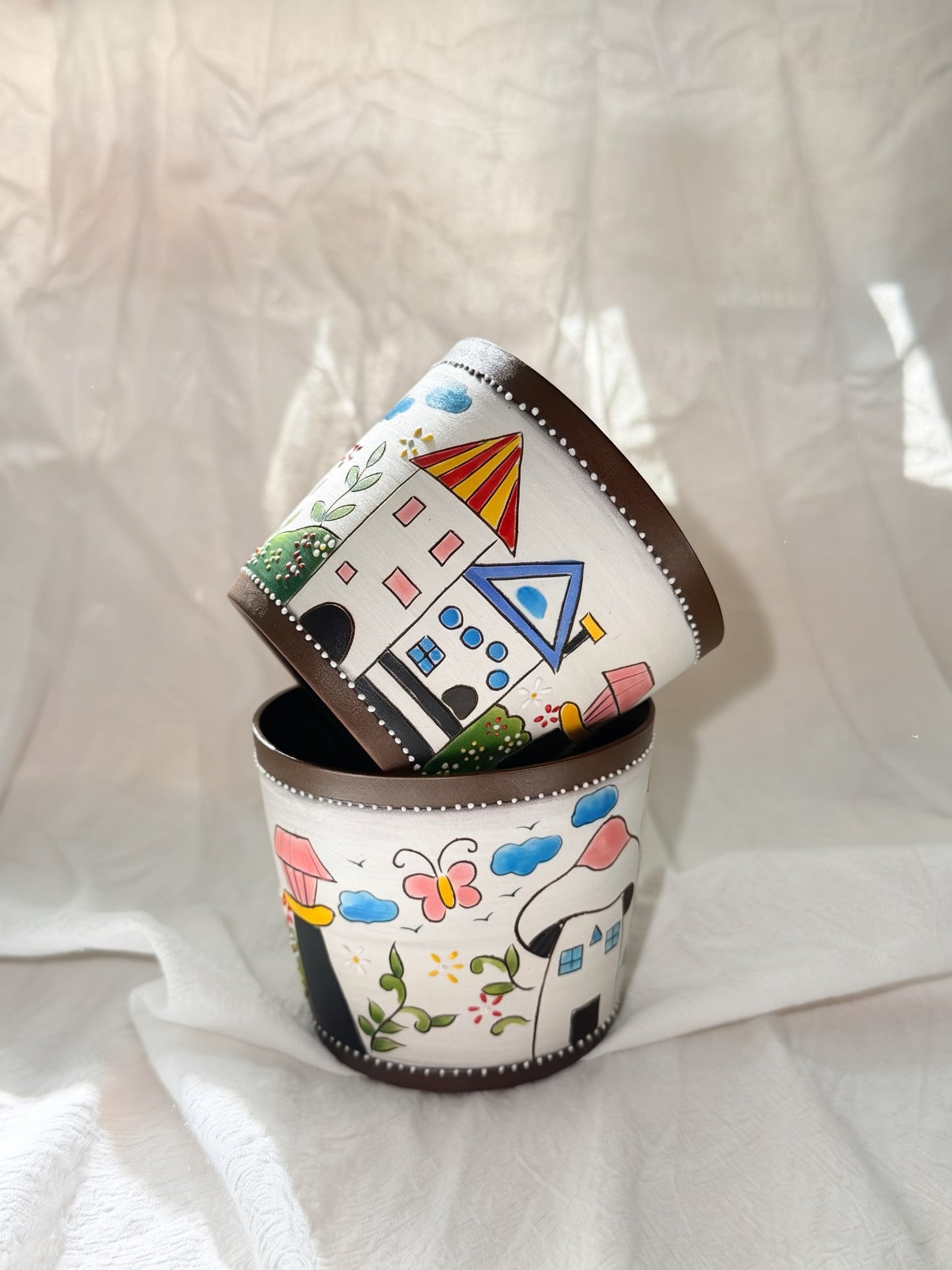 Three small cylindrical white containers with dark brown rims, stacked with one tilted on top of the other. The containers are decorated with colorful hand-painted whimsical illustrations of houses, clouds, butterflies, and flowers, set against a draped white fabric background.