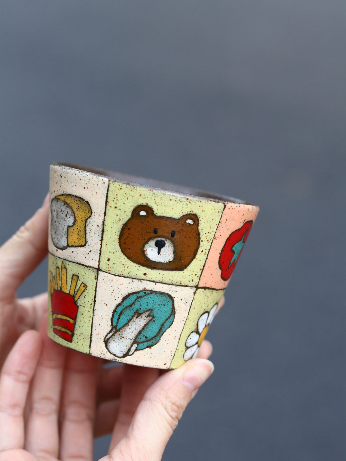 Hand holding a colourful patchwork ceramic cup featuring fruit and dog motifs.