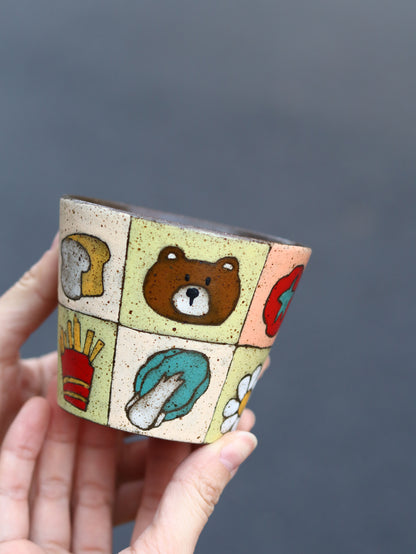 Hand holding a colourful patchwork ceramic cup featuring fruit and dog motifs.