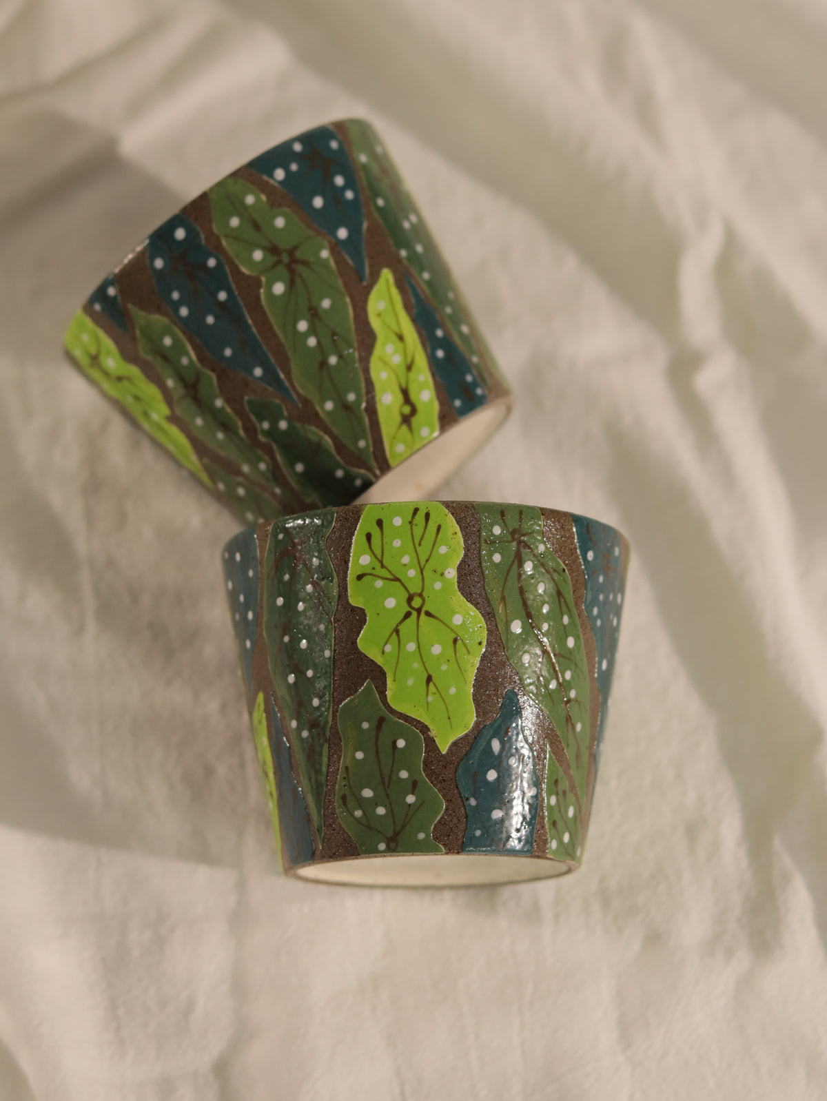 Two hand-carved botanical ceramic cups displayed together on a neutral fabric background.