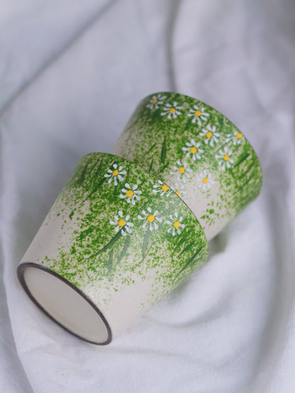 Two ceramic cups with a green grass and daisy pattern, resting on a soft white fabric background.