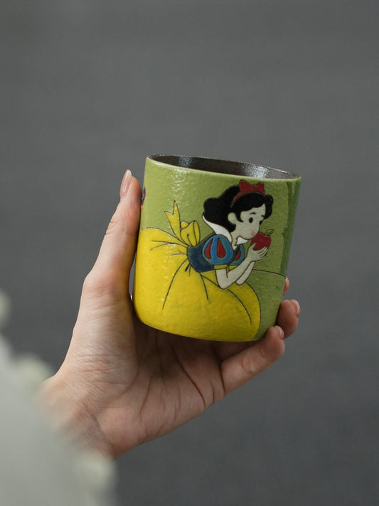 Hand holding a green ceramic cup featuring hand-painted Snow White with the enchanted apple.