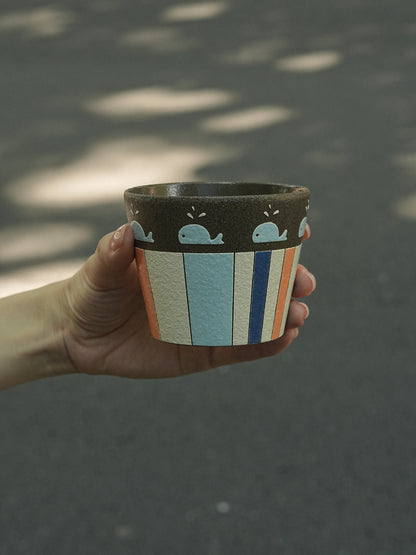 A hand holds a small handmade ceramic planter outdoors against a blurred asphalt background with dappled sunlight. The planter has a dark brown top band with light blue whale illustrations, and a lower body featuring vertical stripes in light blue, orange, white, and dark blue.