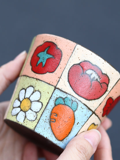 Close-up of a hand-painted patchwork cup showing tomato, flower and carrot illustrations.