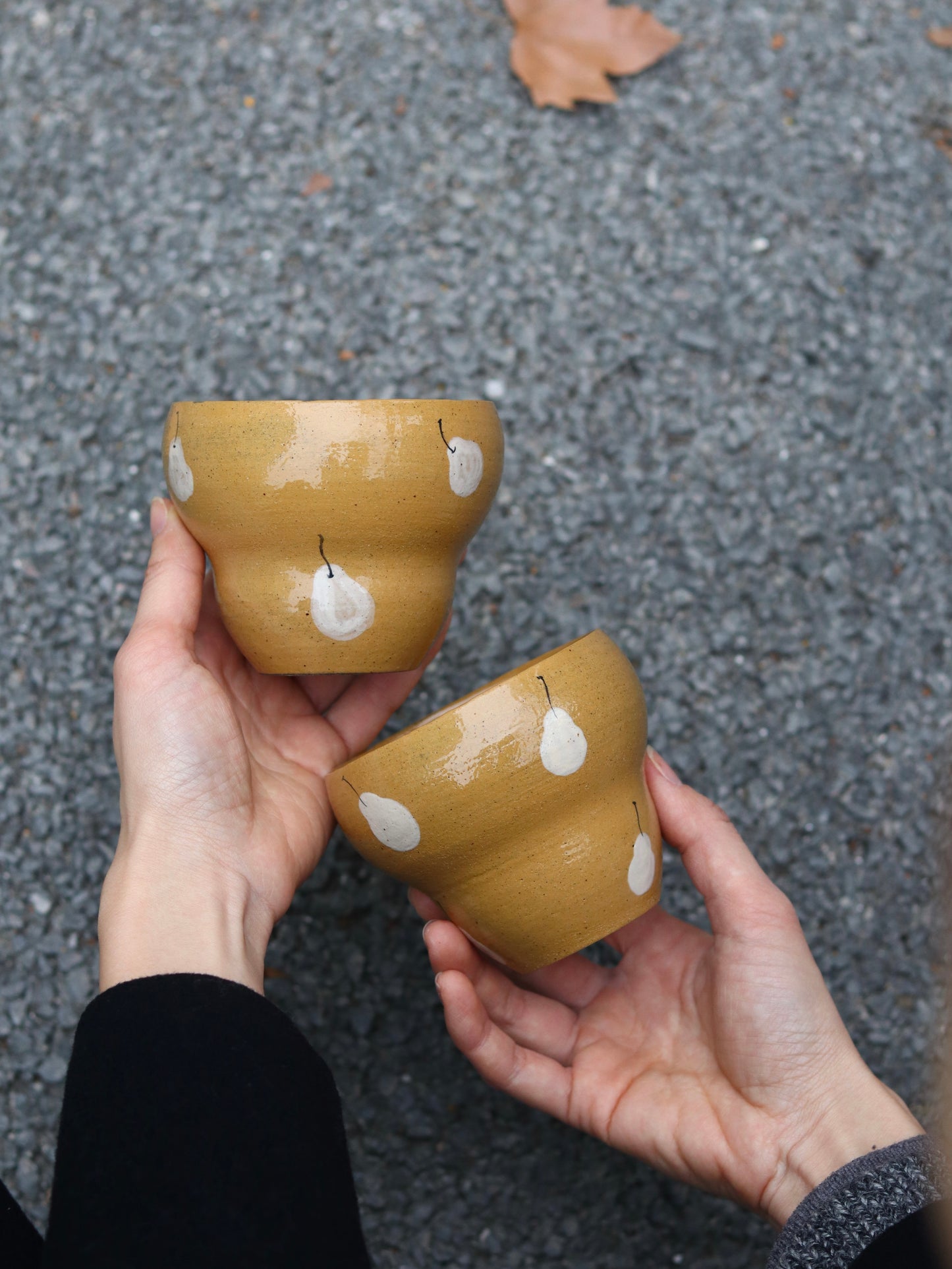 Two hands holding two small ceramic pots with white floral patterns on a gray pavement background.