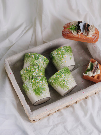 Four ceramic cups decorated with a green grass and white daisy floral pattern, arranged in a rustic woven tray on a white fabric background with decorative pastries.