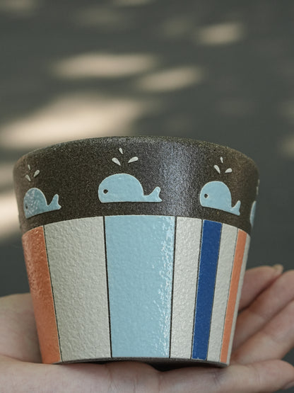A hand holds a small cylindrical cup or planter with a dark brown top band decorated with light blue whale illustrations, and a lower body featuring vertical stripes in cream, light blue, orange, and blue.