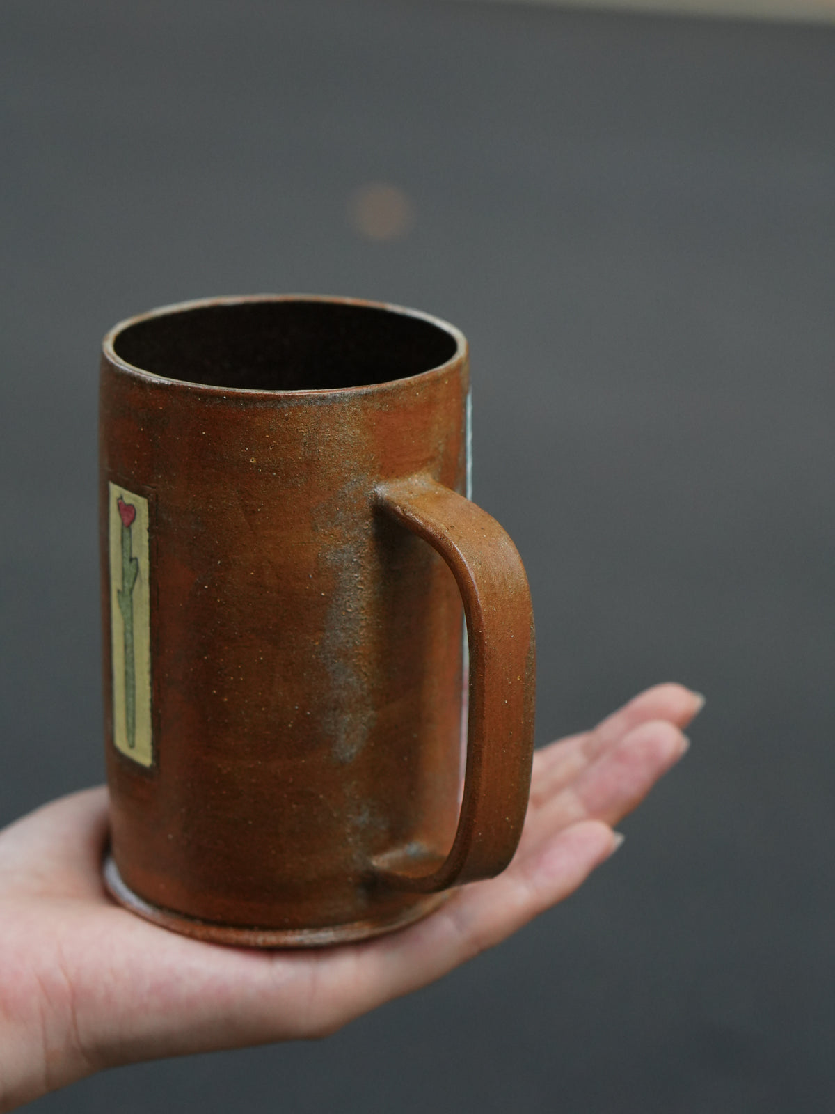 A handmade brown ceramic mug with a handle and a small vertical floral label, held in a person's hand against a dark gray background.