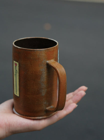 A handmade brown ceramic mug with a handle and a small vertical floral label, held in a person's hand against a dark gray background.