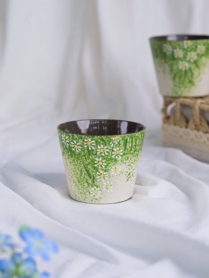 A small ceramic cup with a green and white daisy floral pattern, set on draped white fabric with another matching cup blurred in the background.