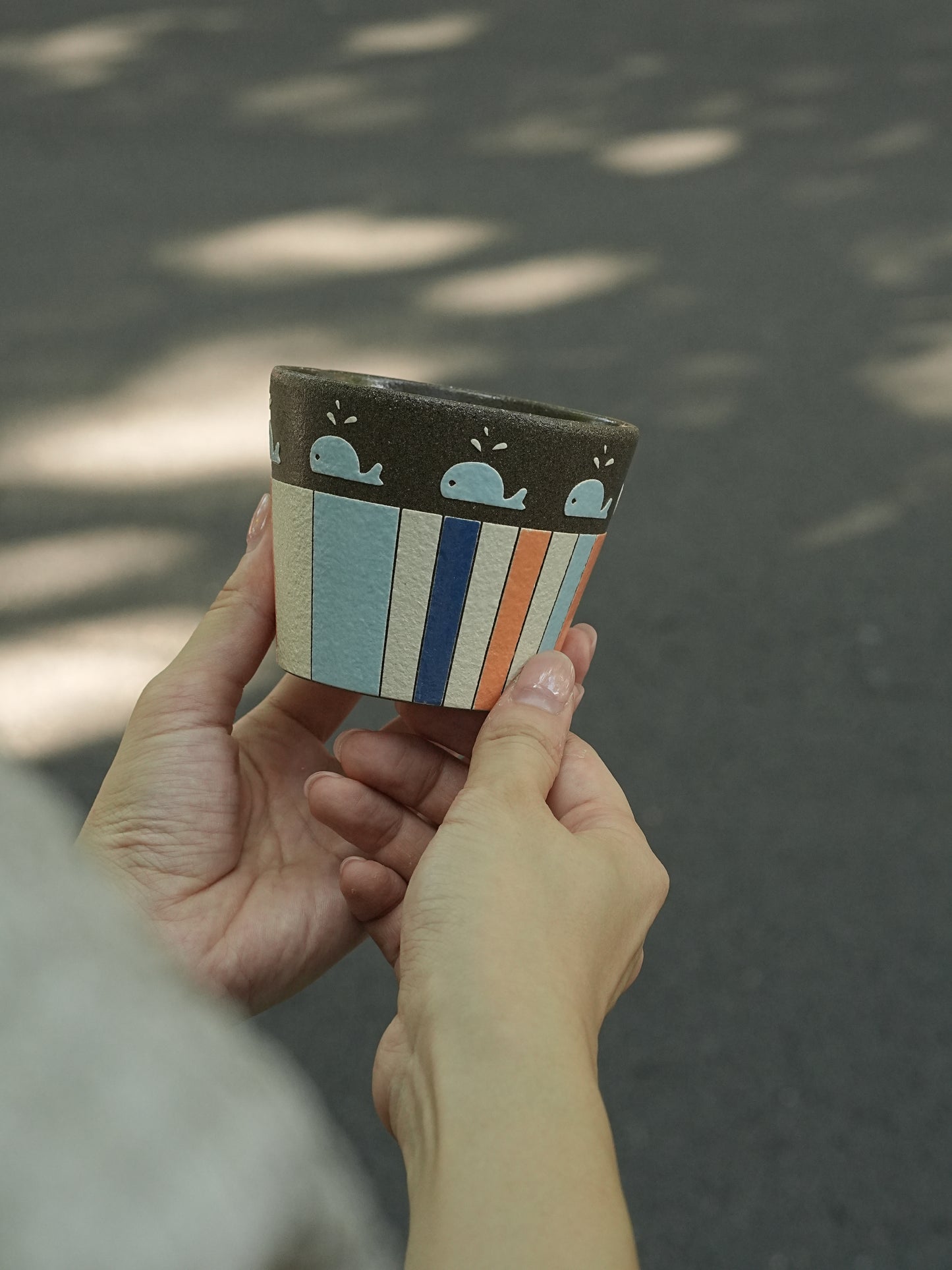 A person holds a small ceramic planter pot outdoors. The pot has a dark brown top band with light blue whale illustrations, and a body with vertical stripes in light blue, navy, white, and orange.