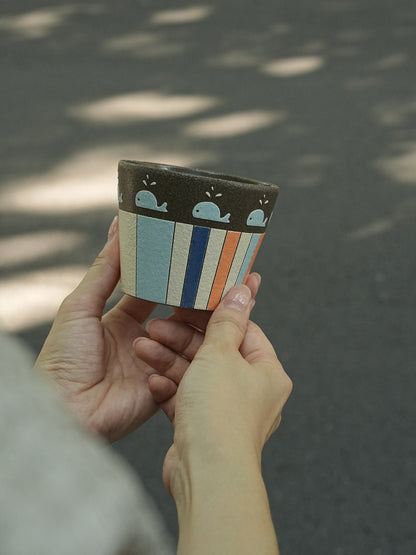 A person holds a small ceramic planter pot outdoors. The pot has a dark brown top band with light blue whale illustrations, and a body with vertical stripes in light blue, navy, white, and orange.