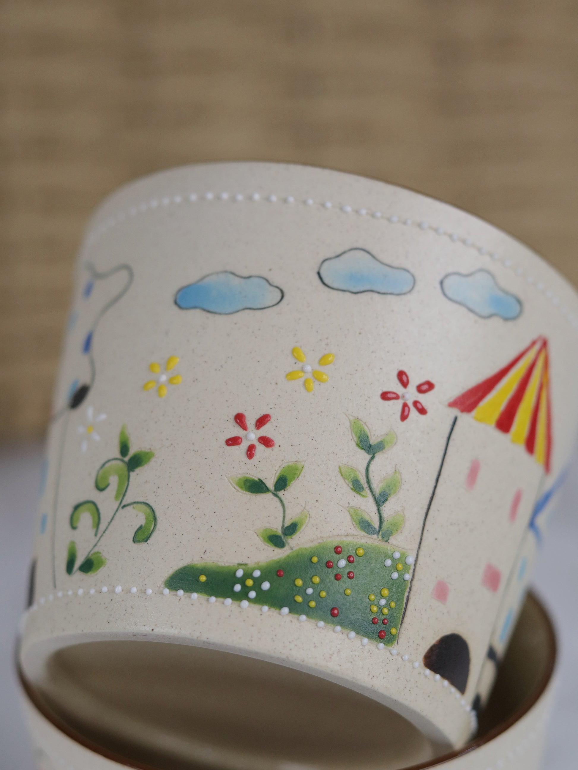Ceramic mug with hand-painted designs of flowers, clouds, and a house on a beige background.