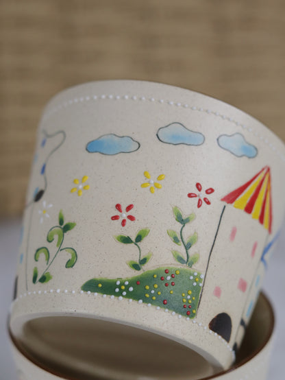 Ceramic mug with hand-painted designs of flowers, clouds, and a house on a beige background.