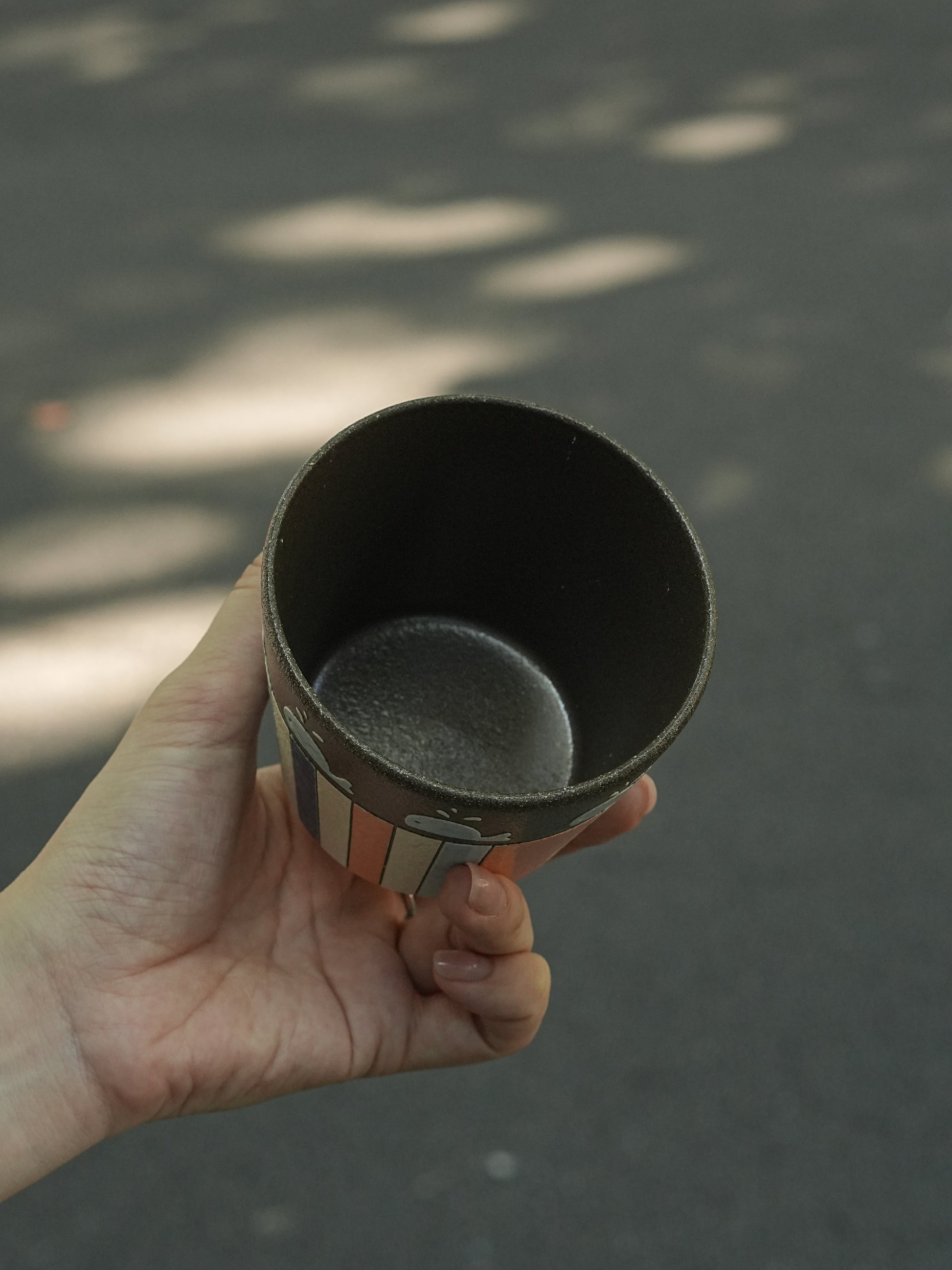 A hand holding a dark ceramic cup outdoors against a blurred asphalt background with dappled sunlight.