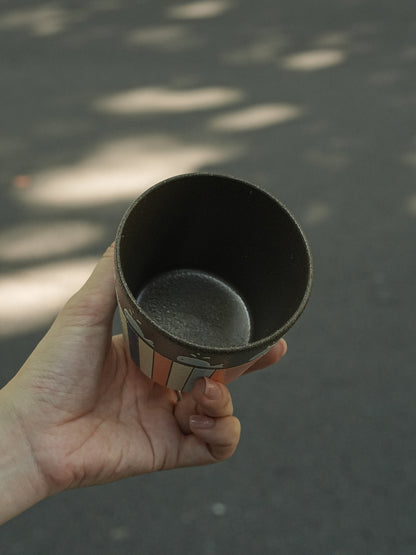 A hand holding a dark ceramic cup outdoors against a blurred asphalt background with dappled sunlight.