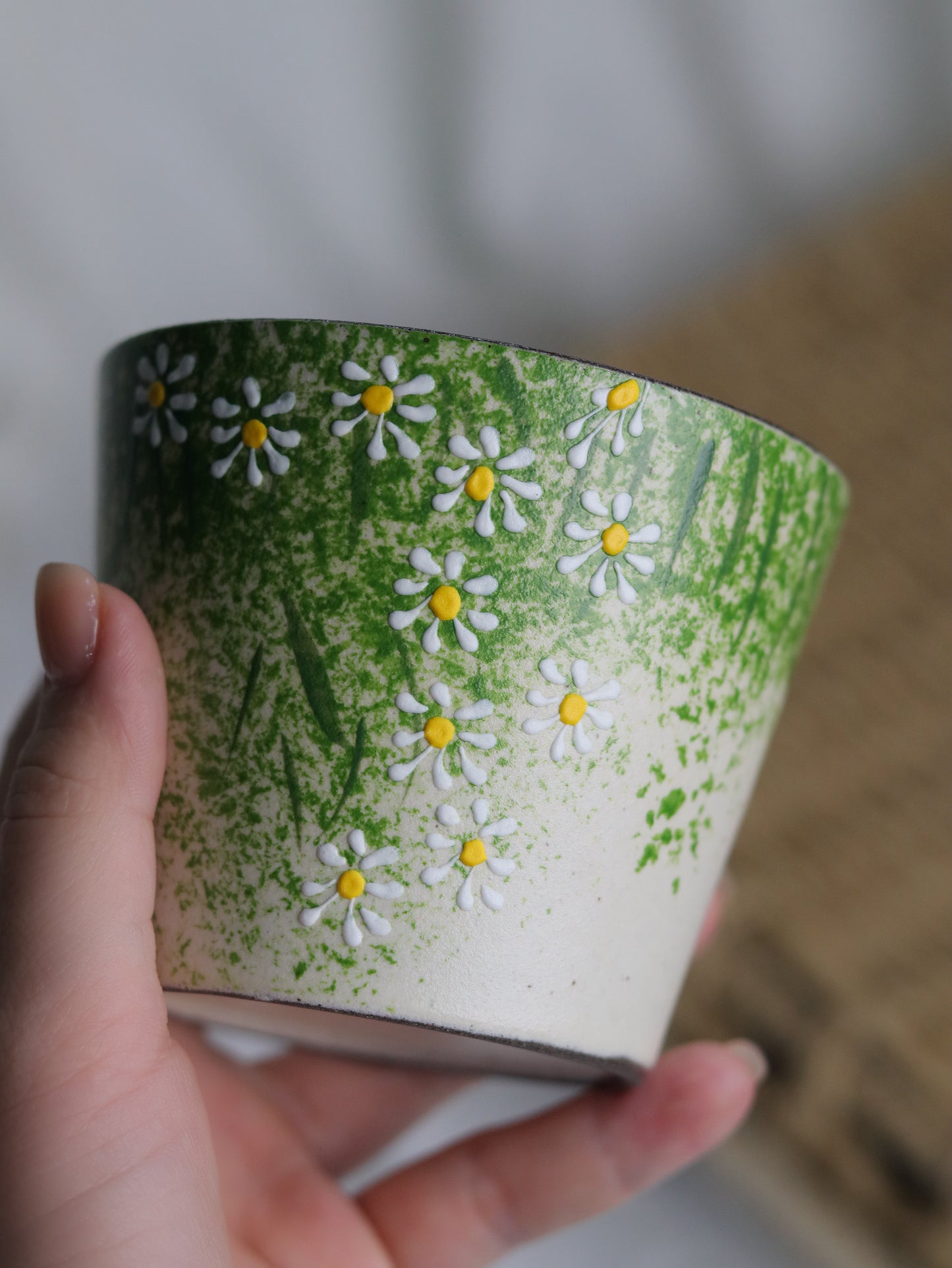 A hand holding a handmade ceramic cup decorated with a green and white daisy pattern, set against a soft-focus background.