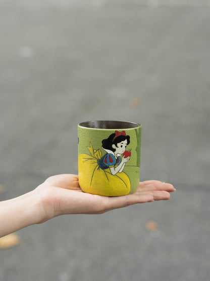 Hand holding a green ceramic cup featuring hand-painted Snow White with the enchanted apple.