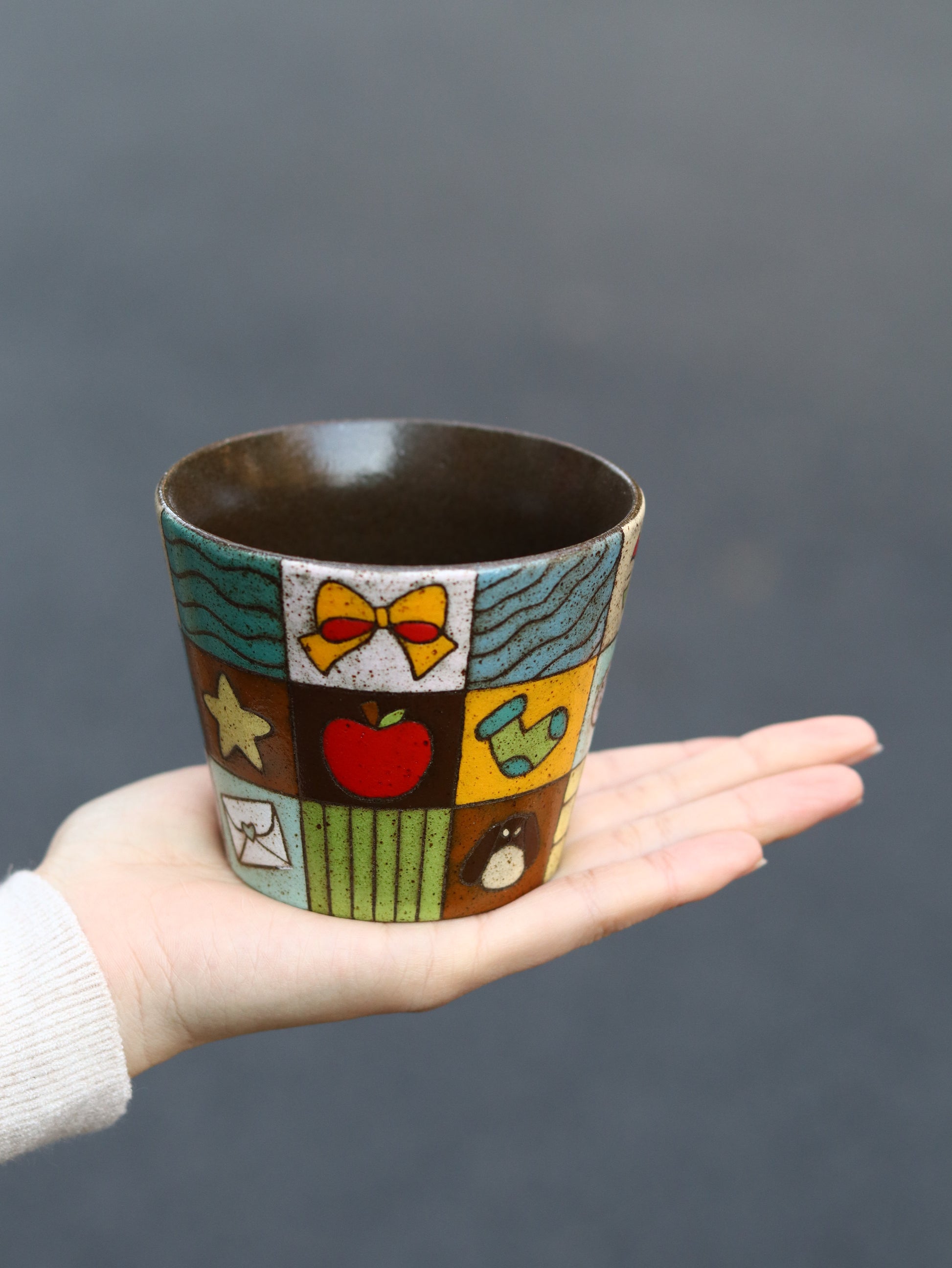 Hand holding a colorful ceramic cup with various designs against a dark background