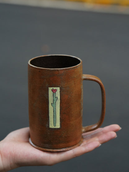 A handmade brown ceramic mug with a handle, held in a person's hand against a blurred gray background. The mug features a vertical rectangular label depicting a green cactus with a small red heart on its tip.
