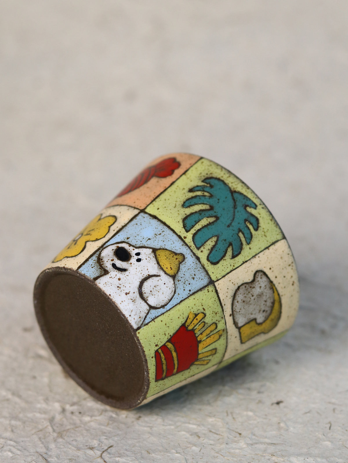 Patchwork ceramic cup resting on its side, showing the colourful wraparound design and pottery clay base.