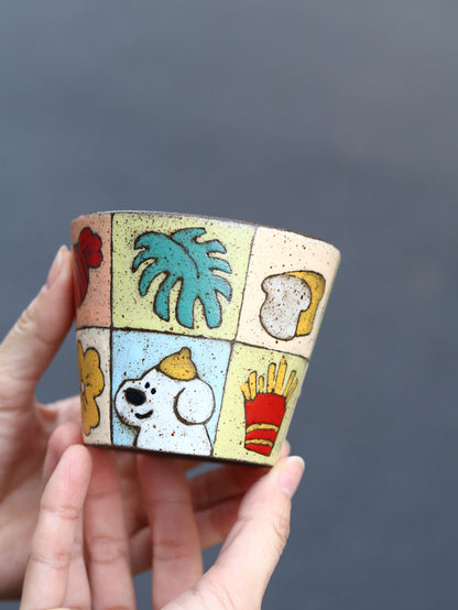 Hand holding a patchwork ceramic cup featuring a playful dog and botanical motifs.
