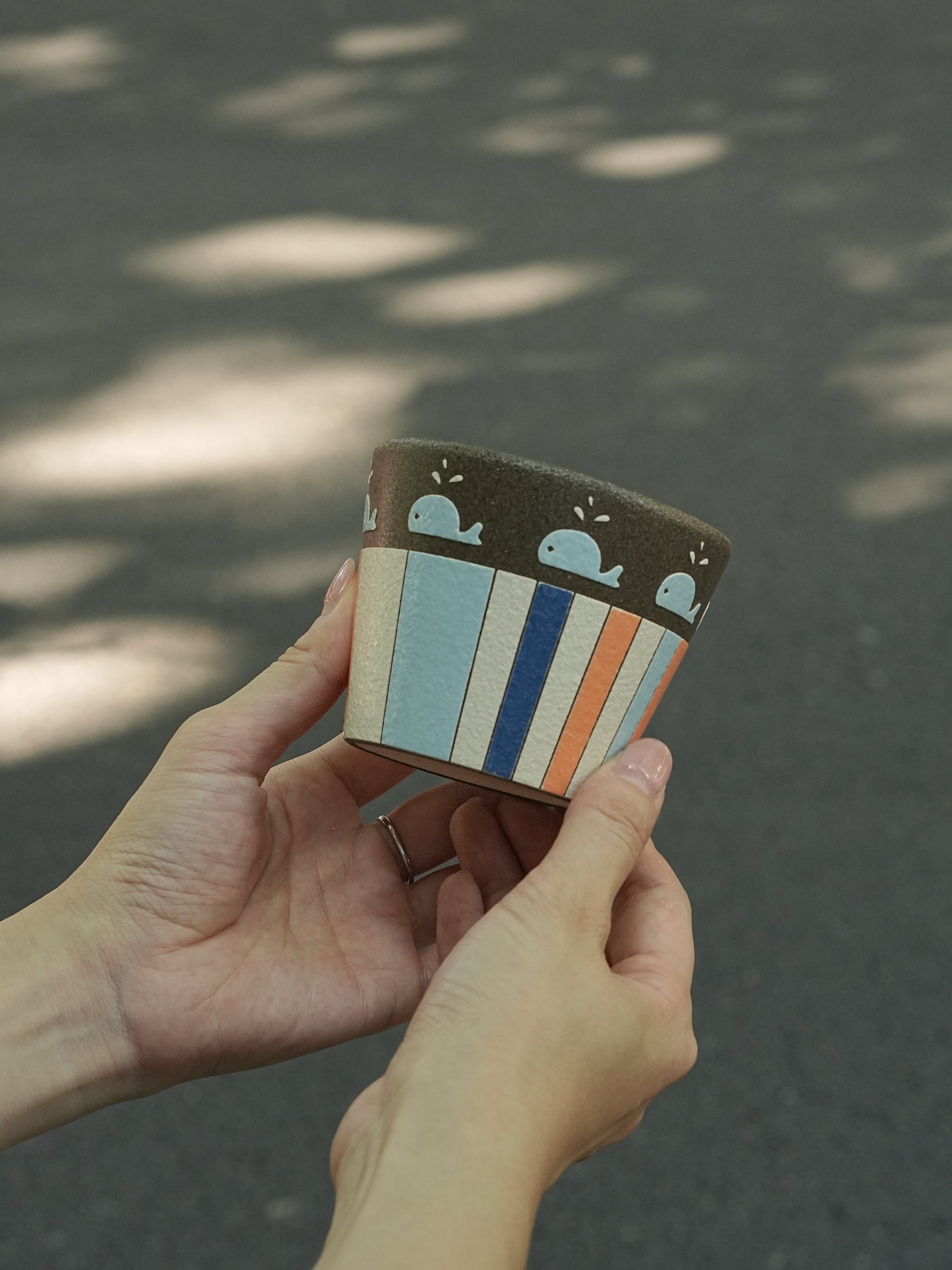 A person's hands holding a small, hand-painted ceramic planter outdoors on a paved surface with dappled sunlight.