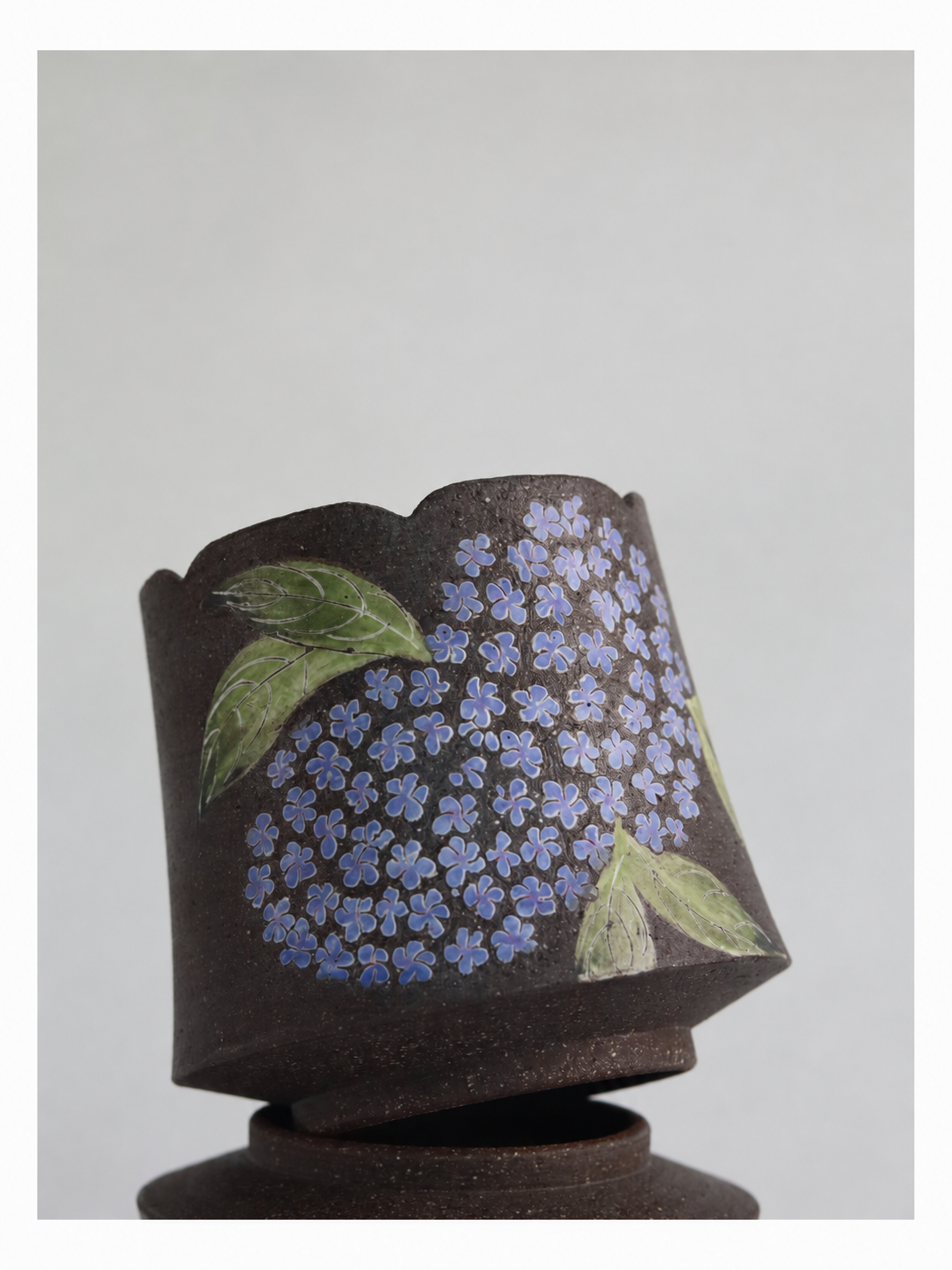 Close-up of a zisha clay cup showing blue hydrangea blossoms, green leaves and a scalloped rim.