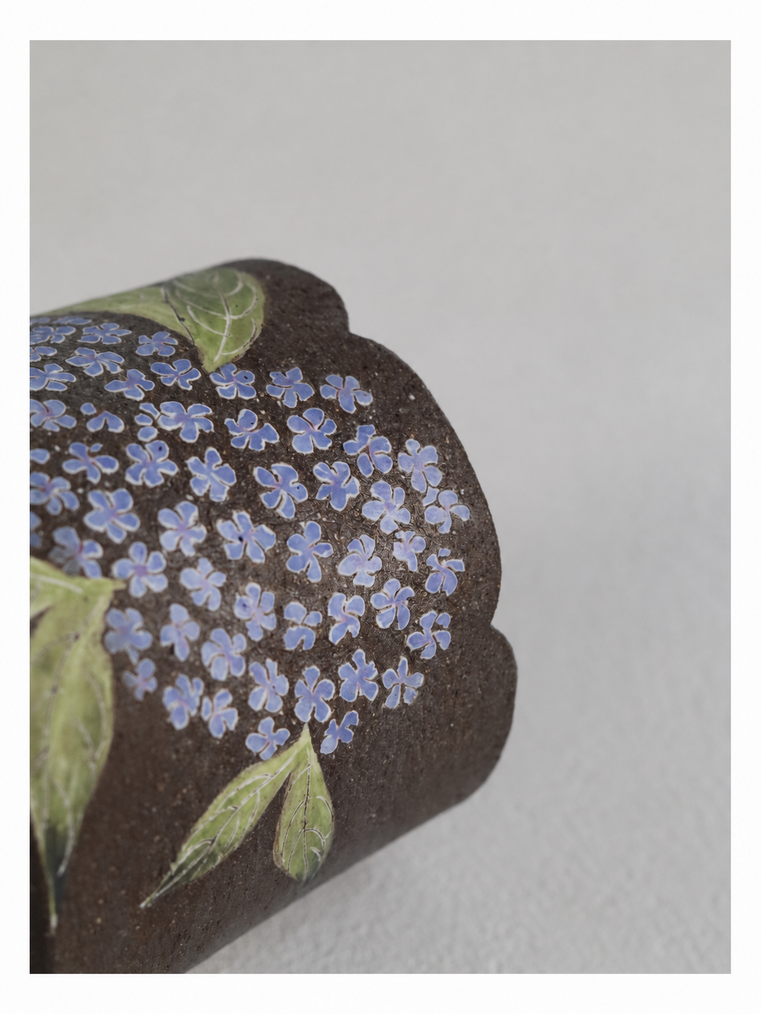 Close-up of a zisha clay cup showing blue hydrangea blossoms, green leaves and a scalloped rim.