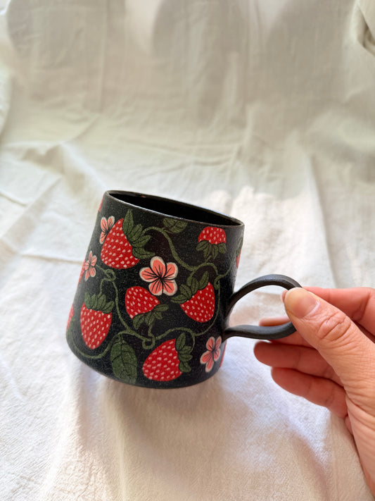 Black mug with strawberry design held by a hand on a white fabric background