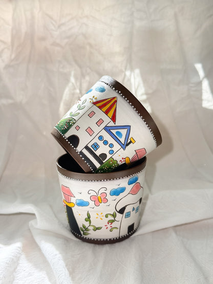Three small cylindrical white containers with dark brown rims, stacked with one tilted on top of the other. The containers are decorated with colorful hand-painted whimsical illustrations of houses, clouds, butterflies, and flowers, set against a draped white fabric background.