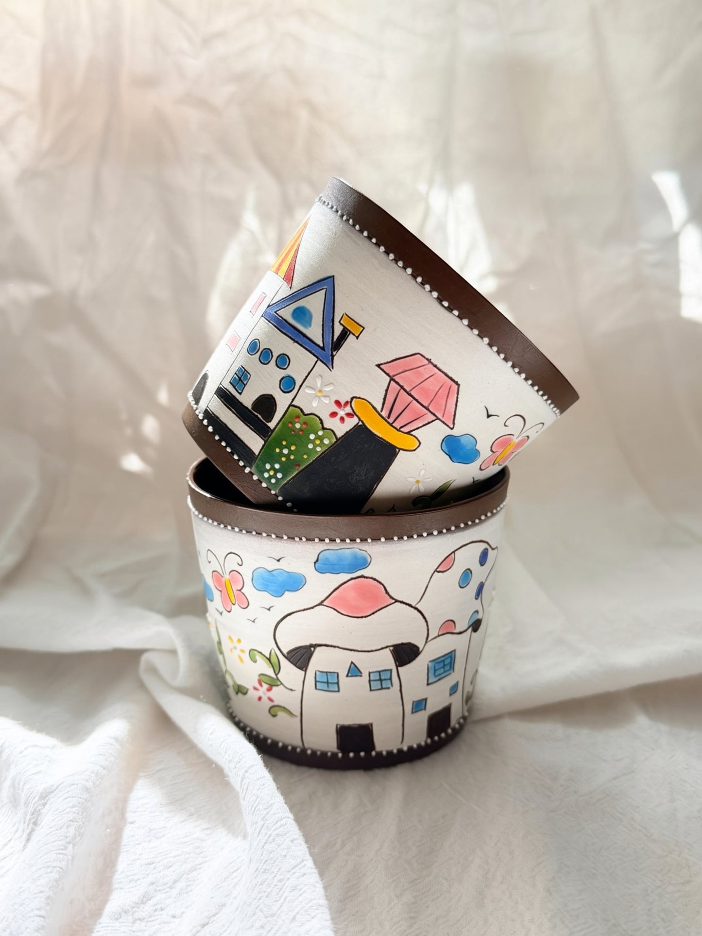 A stack of three hand-painted ceramic cups with whimsical house and cloud illustrations, set against a draped white fabric background.