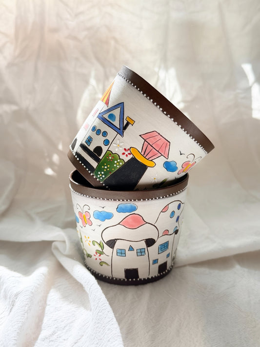 A stack of three hand-painted ceramic cups with whimsical house and cloud illustrations, set against a draped white fabric background.