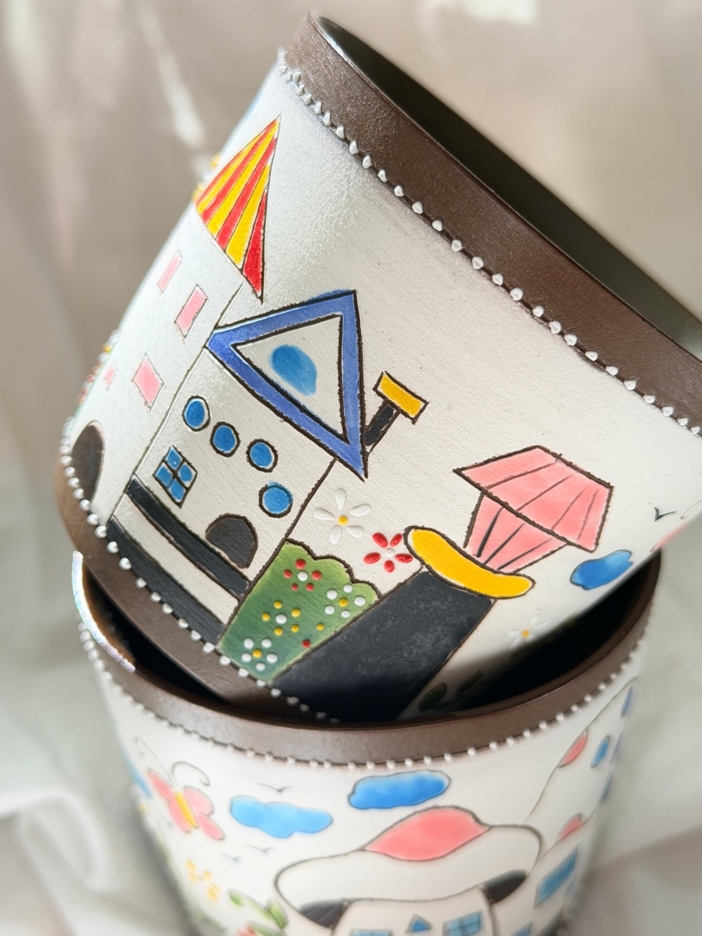 A close-up of stacked cylindrical containers or cups with a white base, brown rim, and colorful hand-painted whimsical house and cloud designs.