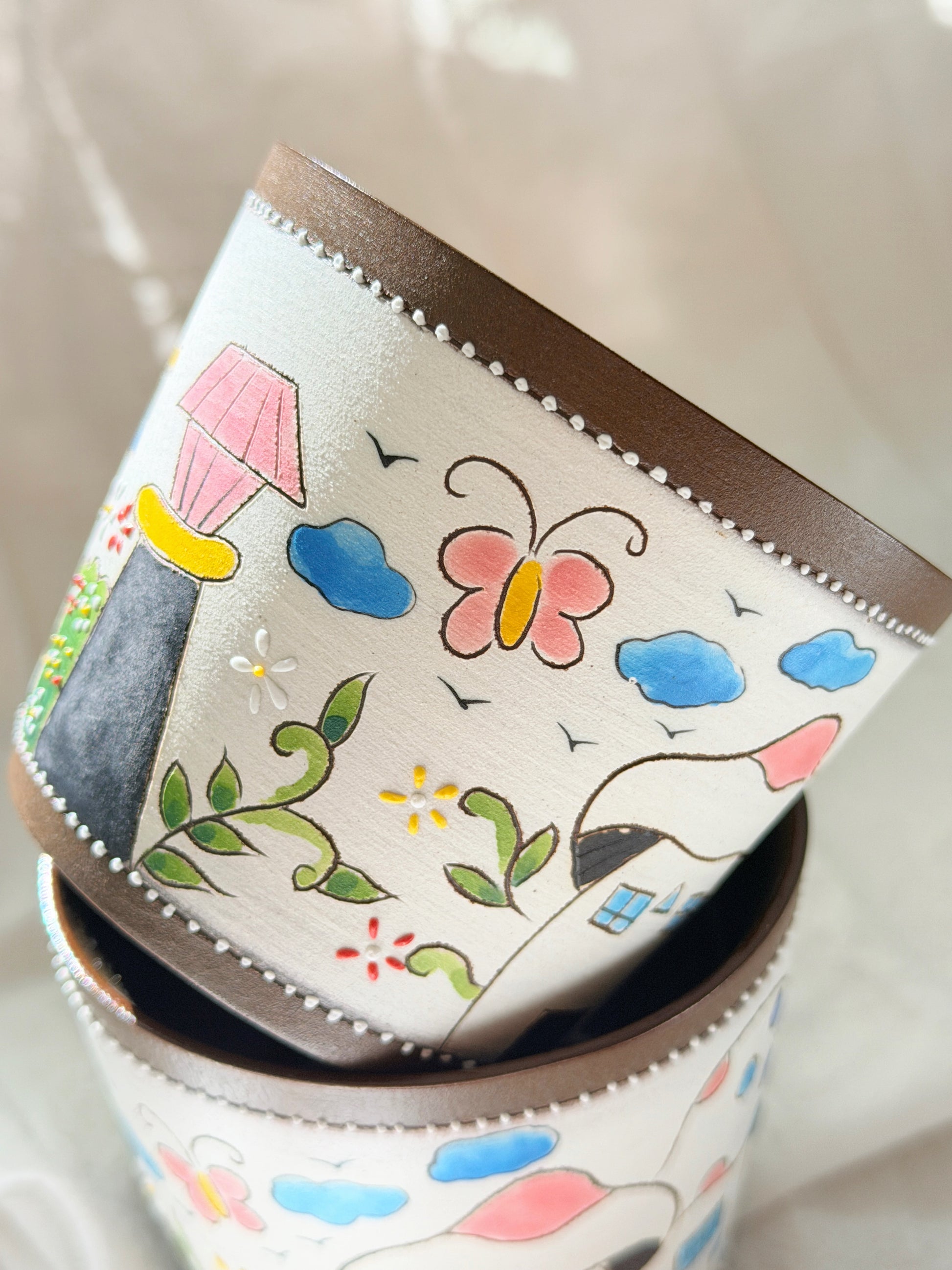 A close-up of a cylindrical container or cup with a cream-colored surface decorated with colorful hand-painted style illustrations, including a butterfly, clouds, flowers, birds, and a small house. The rim is dark brown with a row of small white dots around the edge.
