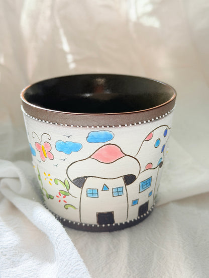 A white ceramic cup decorated with a whimsical hand-painted scene of mushroom houses, blue clouds, a butterfly, and flowers, with a dark brown interior and rim.