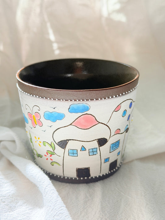 A white ceramic cup decorated with a whimsical hand-painted scene of mushroom houses, blue clouds, a butterfly, and flowers, with a dark brown interior and rim.