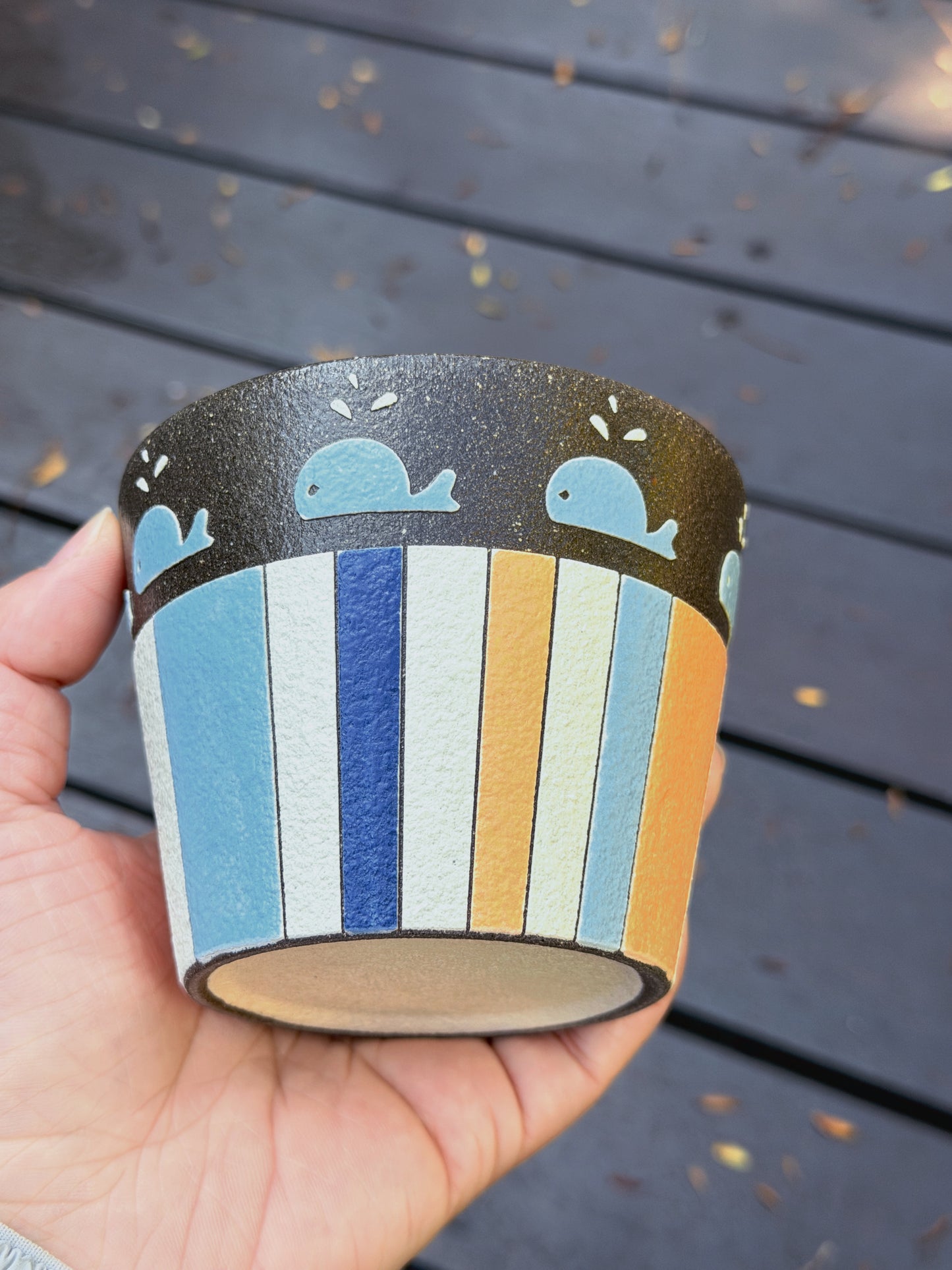 A hand holding a small cylindrical cup or tumbler with a dark top band decorated with light blue whale illustrations, and a lower body featuring vertical stripes in light blue, navy, white, beige, and mustard yellow.