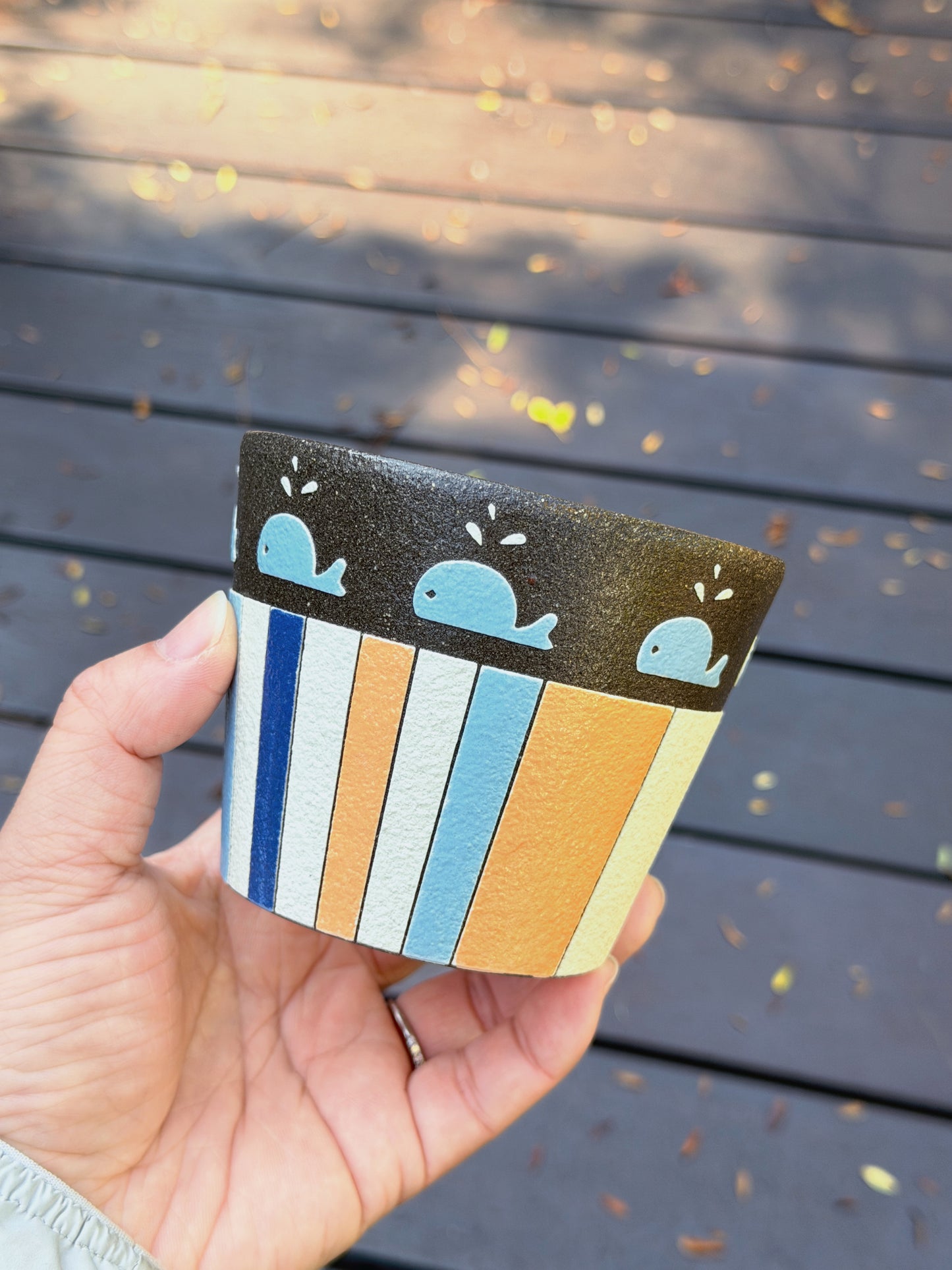 A hand holding a small decorative cup outdoors against a dark wooden deck background. The cup has a black top band with three light blue whale illustrations, and vertical stripes below in navy, white, light blue, and mustard yellow.