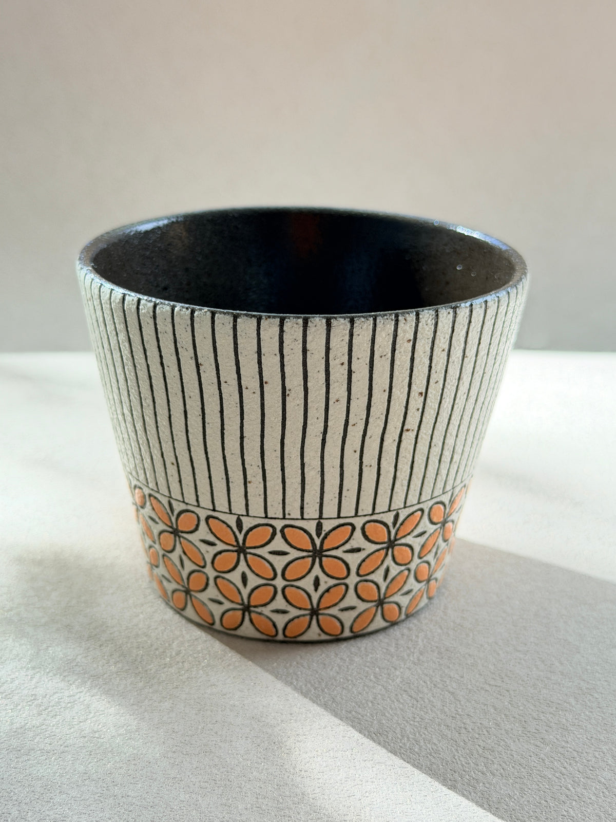 A handmade ceramic cup with a speckled beige exterior, featuring vertical black stripes on the upper half and a lower band of orange and black floral motifs, set against a neutral light gray background.