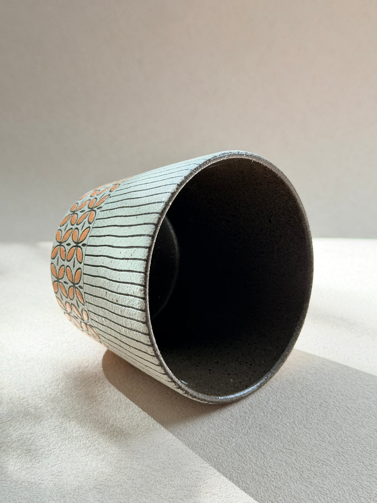 A handmade ceramic cup with a dark speckled interior and exterior featuring a white base with thin vertical gray lines and a peach-colored floral pattern near the rim, set against a neutral beige background.