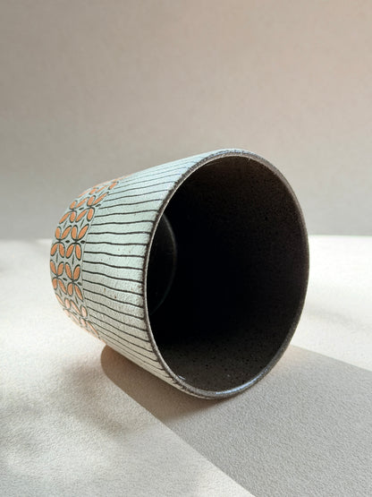 A handmade ceramic cup with a dark speckled interior and exterior featuring a white base with thin vertical gray lines and a peach-colored floral pattern near the rim, set against a neutral beige background.