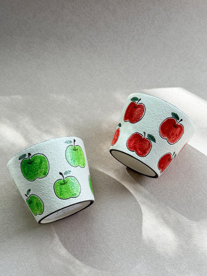 Red and green apple ceramic cups resting on their sides in natural sunlight.