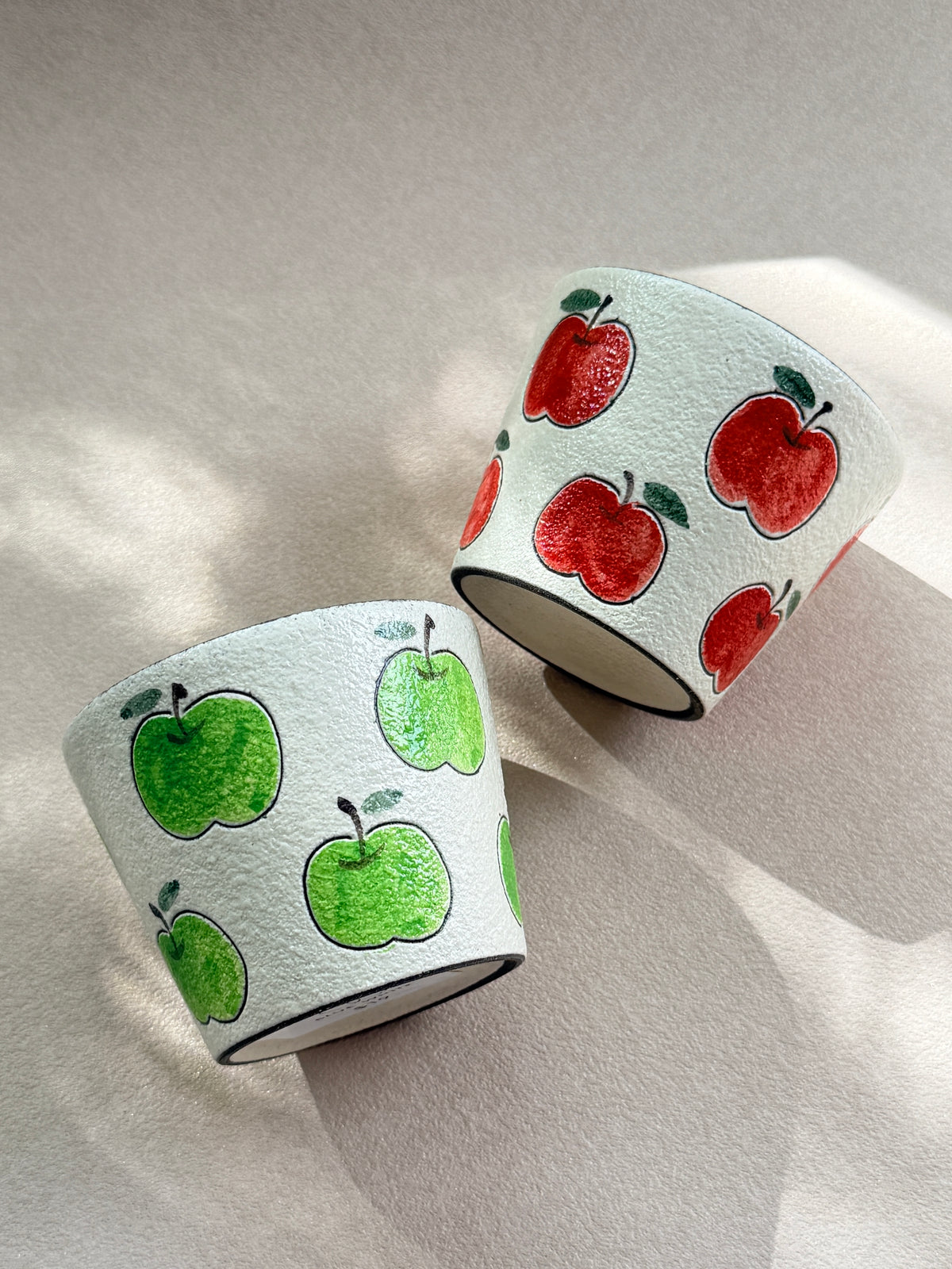 Red and green hand-painted apple ceramic cups with textured ivory finishes and dark glazed interiors.