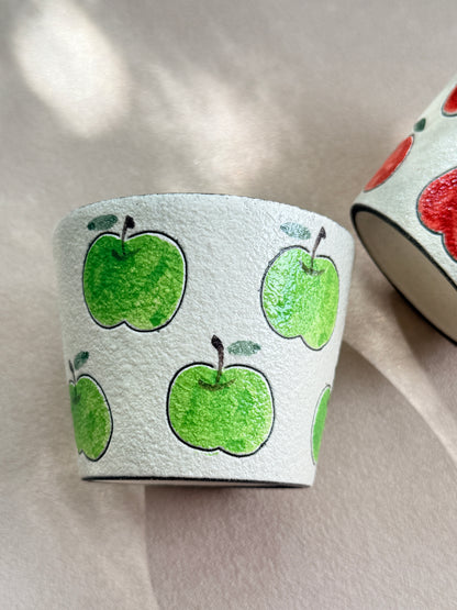 A white textured leather cup decorated with hand-painted green apples, shown alongside another cup displaying red apple illustrations on a neutral beige background.