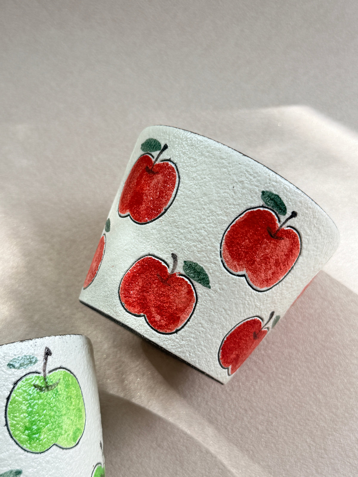 Hand-painted red apple ceramic cup resting on its side beside a green apple cup.