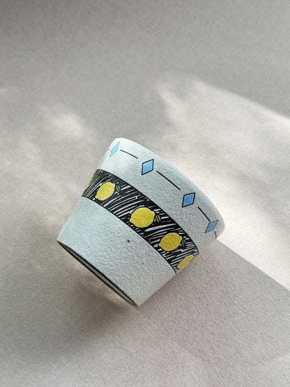 Angled view of the textured lemon ceramic cup in natural sunlight, showing its yellow fruit and pale blue diamond details.
