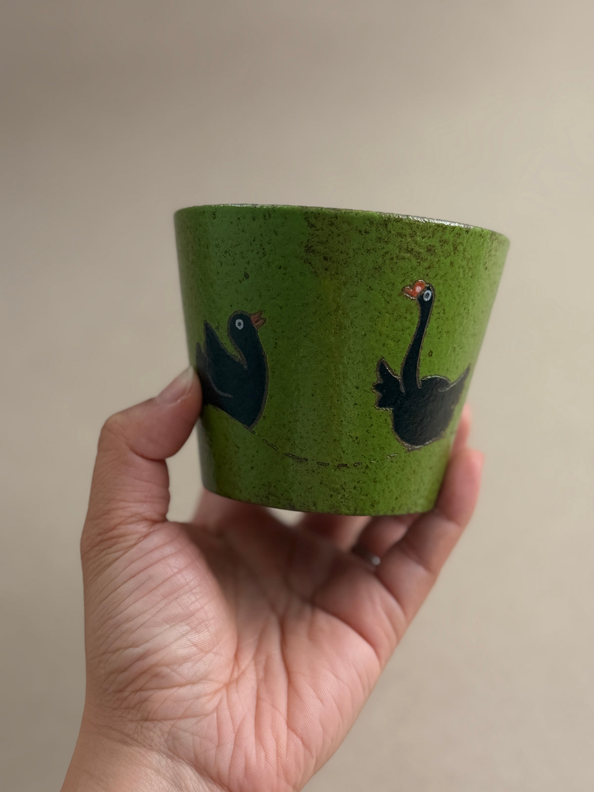 A hand holding a speckled olive green ceramic cup decorated with two black swans, set against a plain beige background.