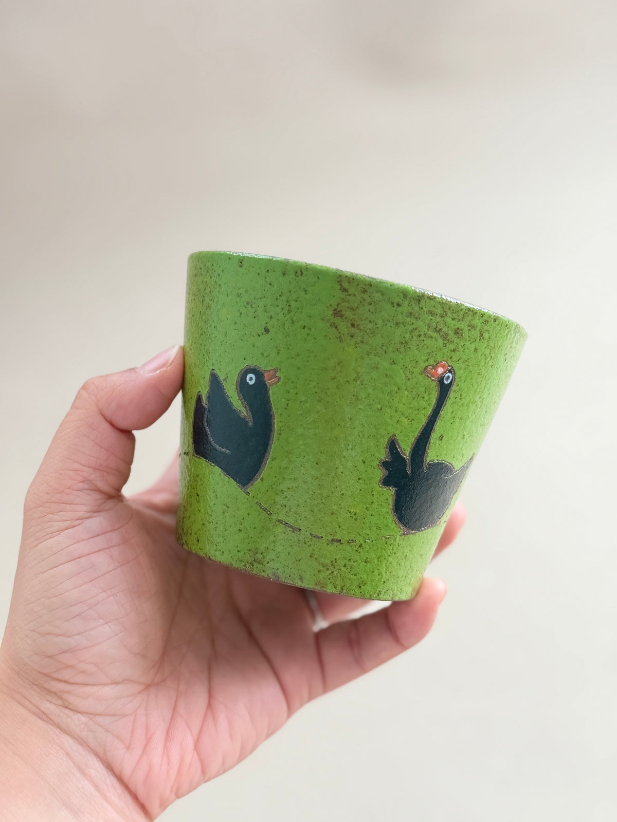 A hand holding a small green speckled ceramic cup decorated with two black birds, one with a red beak and one with a red wattle, against a soft beige background.