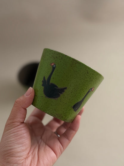 A hand holding a small green ceramic cup decorated with black swan illustrations, set against a soft, out-of-focus beige background.